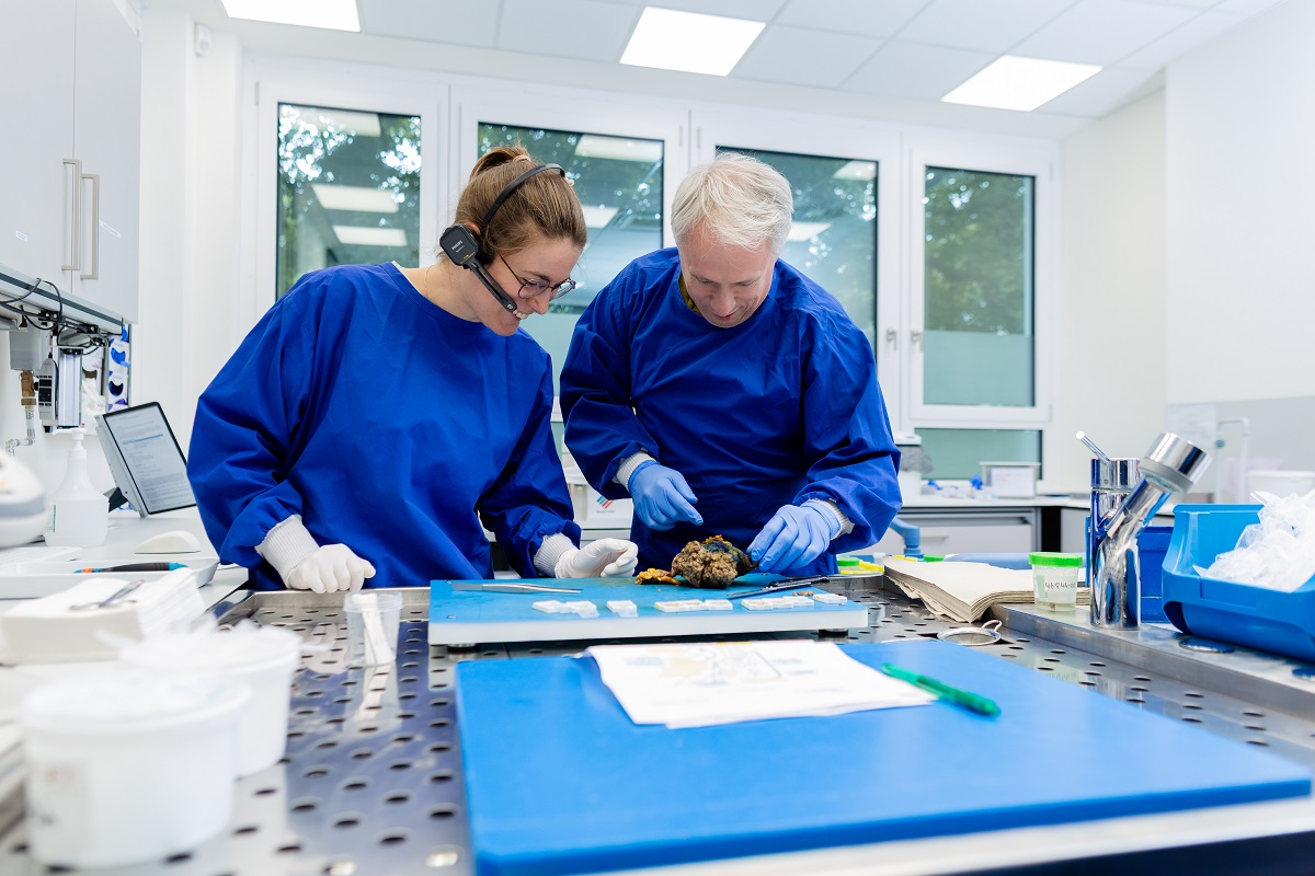 Zwei Mitarbeiterinnen eines Pathologielabors sitzen gegenüber am Labortisch und lächeln